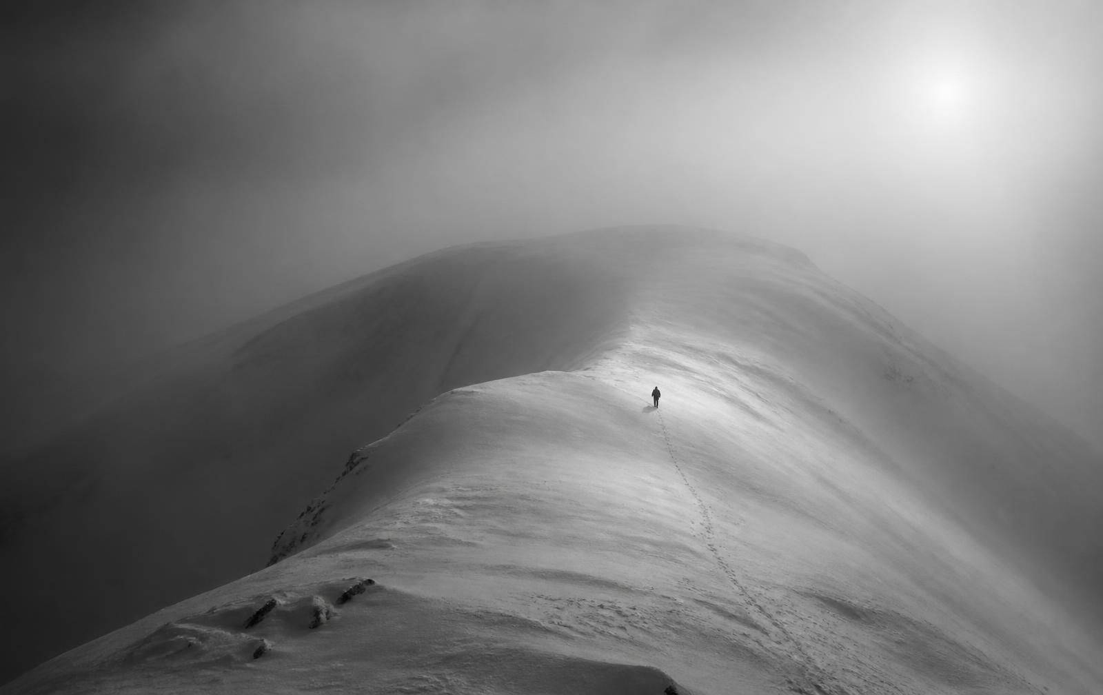 A lone figure walking a vast snow ridge in fog
